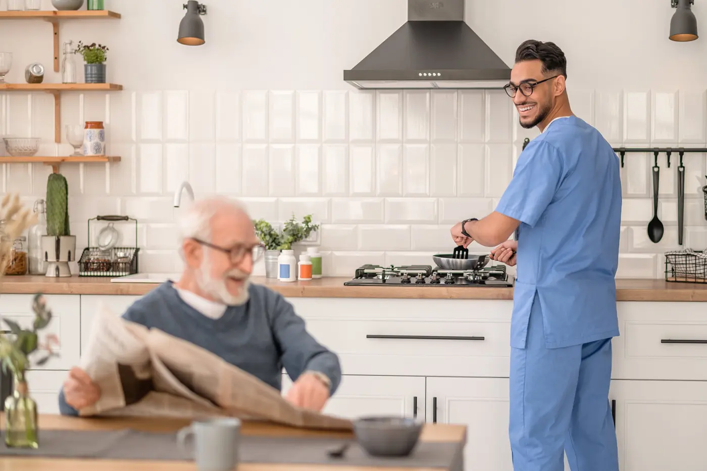 A caregiver cooks breakfast while an older man reads the newspaper