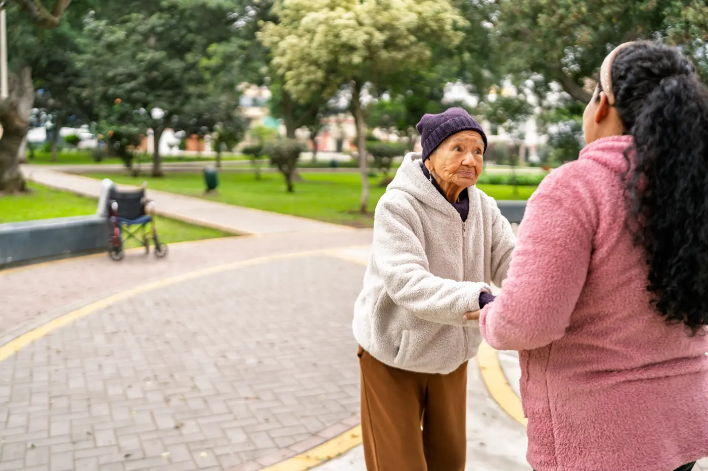 A caregiver holds an older woman's hands as they walk together in a park
